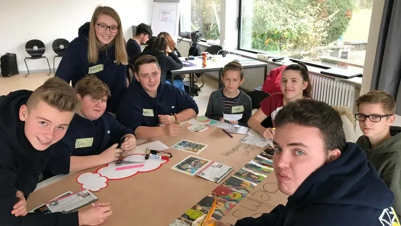 Schülervertretung mit Namensschildern arbeitet an einem Tisch mit Plakat, Bildern und Stiften