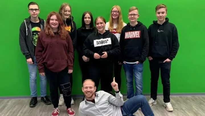 Gruppe von Jugendlichen vor einer grünen Wand, vorne liegt ein Erwachsener und zeigt nach oben