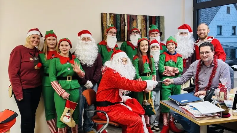 Gruppe in Weihnachtsmann- und Wichtelkostümen in einem Büro, ein Weihnachtsmann reicht einem Mann am Schreibtisch etwas