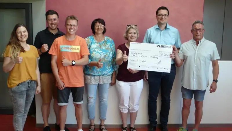 Sieben Erwachsene mit erhobenen Daumen halten einen Spendenscheck über 10.000 Euro