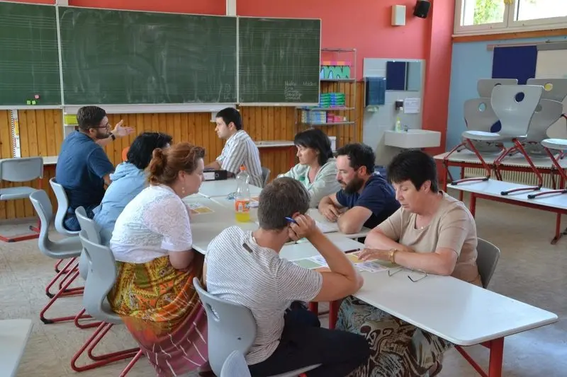 Lehrkräfte sitzen an zusammengestellten Tischen vor einer Tafel und besprechen Arbeitsblätter