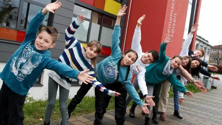 Kinder breiten vor dem Schulgebäude die Arme aus und lächeln in die Kamera