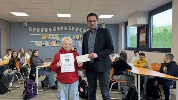 Schüler im roten Pullover hält Urkunde neben einem Erwachsenen mit Umschlag im Klassenraum