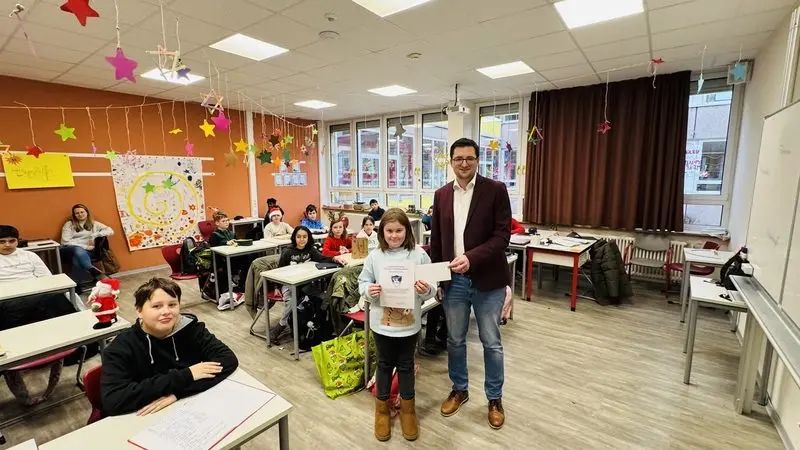 Schülerin mit Urkunde neben einem Mann im weihnachtlich geschmückten Klassenraum, die Klasse sitzt dahinter