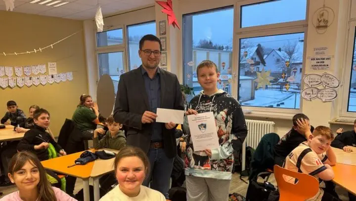 Schüler mit Urkunde und Erwachsener mit Umschlag im Klassenraum, Mitschülerinnen und Mitschüler sitzen an Tischen