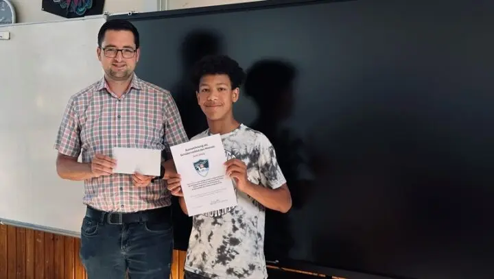 Schüler hält Urkunde neben einem Erwachsenen mit Umschlag vor einer Tafel