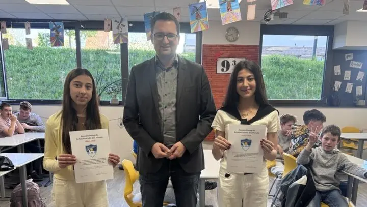 Zwei Schülerinnen halten Urkunden, zwischen ihnen ein Erwachsener im Klassenraum