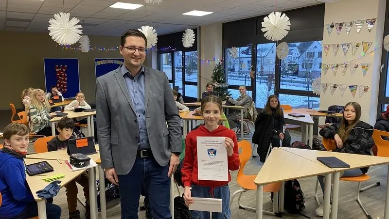 Schülerin mit Urkunde und ein Erwachsener stehen im weihnachtlich geschmückten Klassenraum