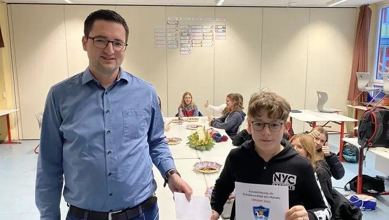 Mann überreicht einem Schüler die Urkunde „Auszeichnung als Schülervorbild des Monats Oktober 2023“, dahinter die Klasse