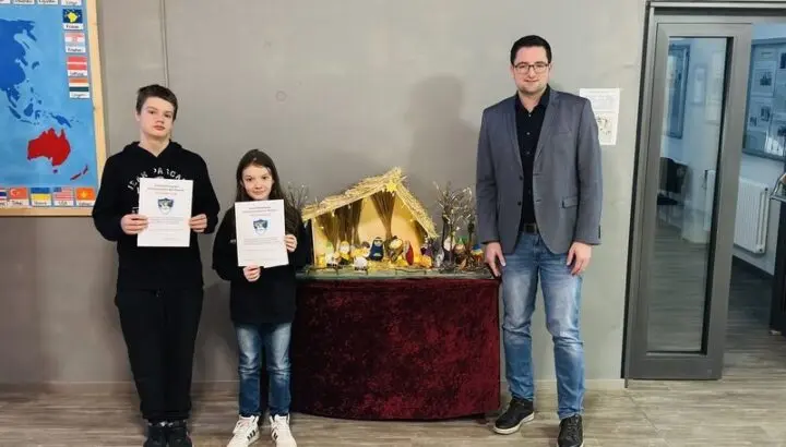 Zwei Jugendliche mit Urkunden und ein Erwachsener neben einer Weihnachtskrippe im Schulfoyer