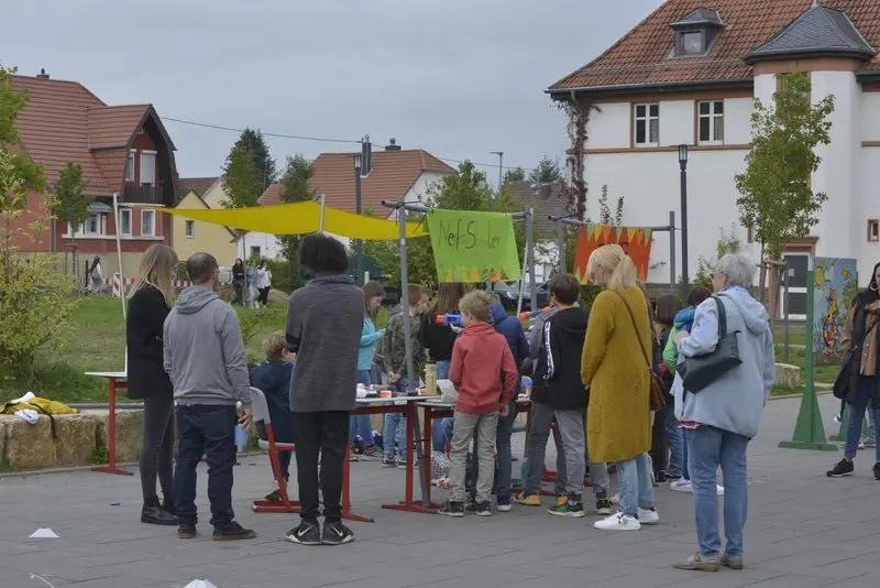 Besucher am Spielstand „Nerf-Shooter“ unter Sonnensegeln auf dem Schulhof beim Schulfest