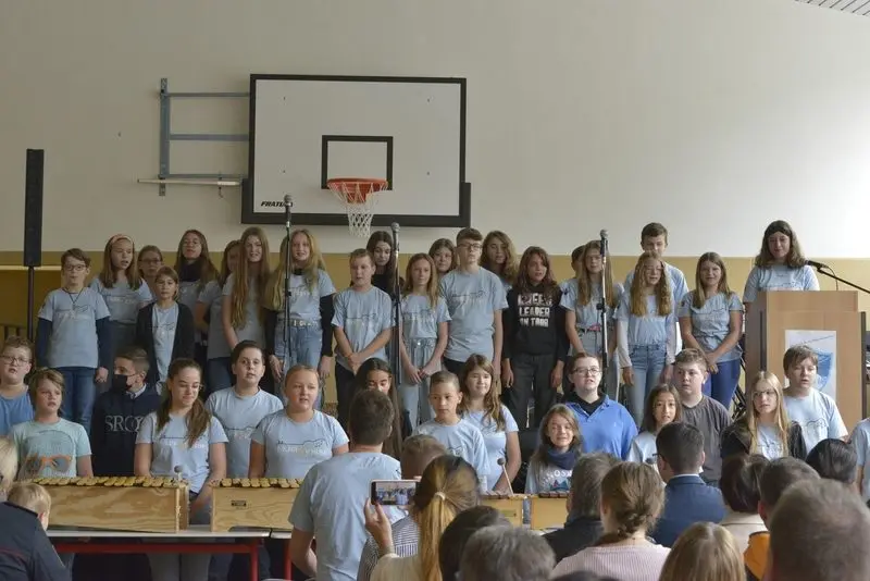 Großer Chor in hellblauen T-Shirts singt in der Sporthalle, davor Xylofone und Publikum