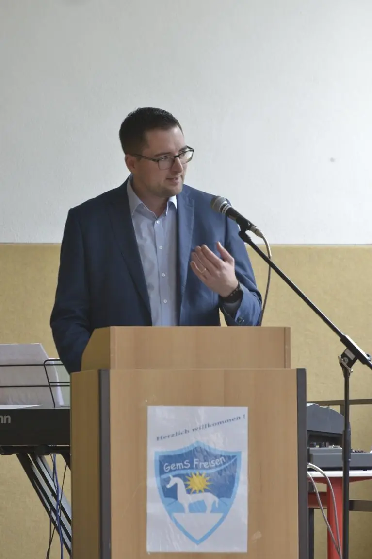 Mann spricht am Rednerpult mit Schulwappen und „Herzlich willkommen!“
