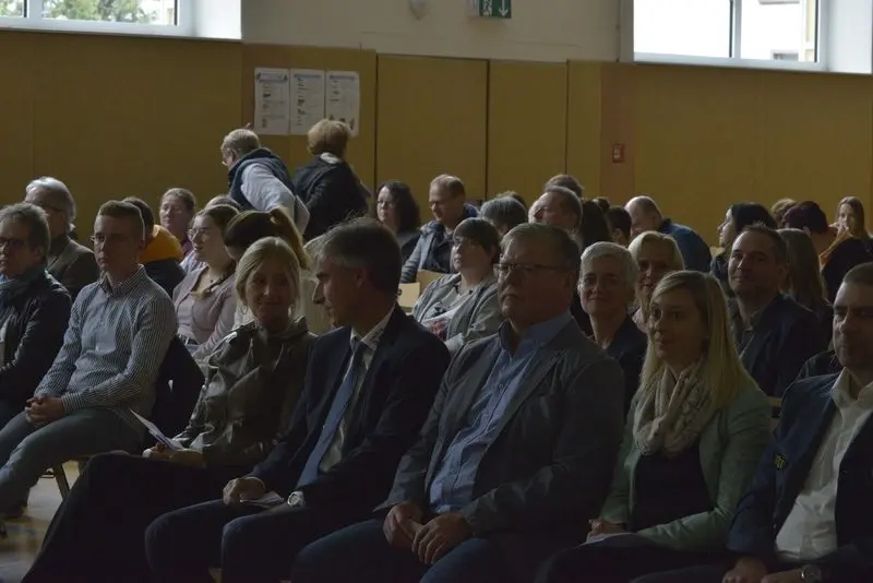 Erwachsene sitzen in Stuhlreihen als Publikum in der Turnhalle
