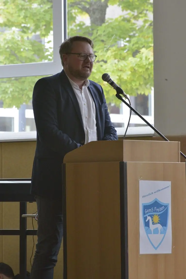Mann spricht am Rednerpult mit Schullogo und „Herzlich willkommen!“-Schild