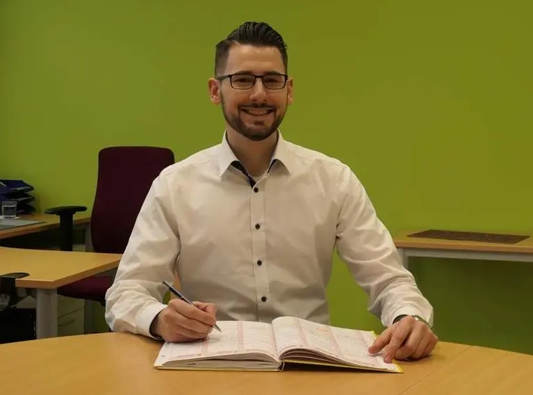 Ein Mann mit Brille sitzt lächelnd am Schreibtisch und schreibt in einen Kalender