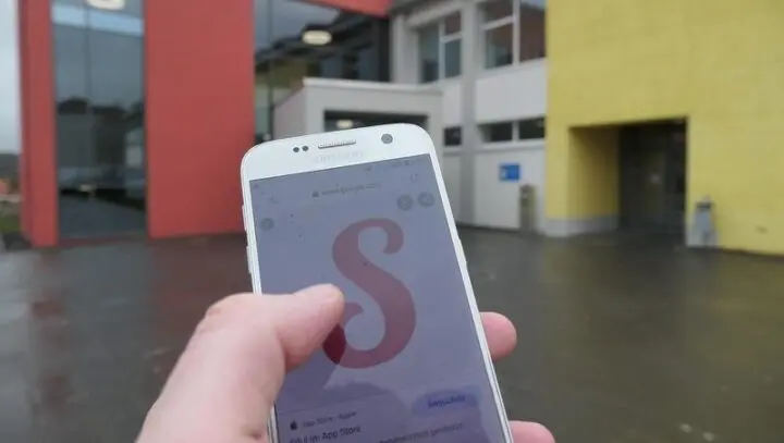 Hand hält Smartphone mit Sdui-Logo vor dem Schulgebäude