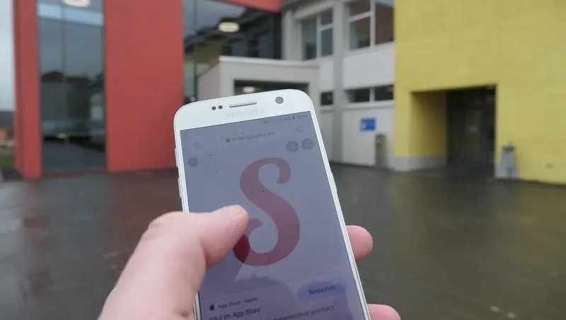 Hand hält Smartphone mit Sdui-Logo vor dem Schulgebäude