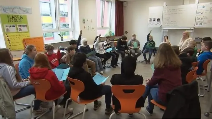 Schülerinnen und Schüler sitzen im Stuhlkreis mit Arbeitsblättern, einige melden sich, Standbild aus einem Fernsehbericht
