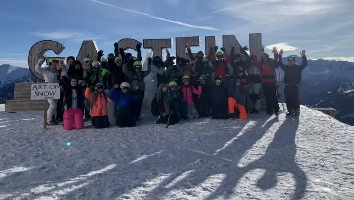 Große Skigruppe jubelt mit erhobenen Armen vor riesigen Holzbuchstaben im Schnee