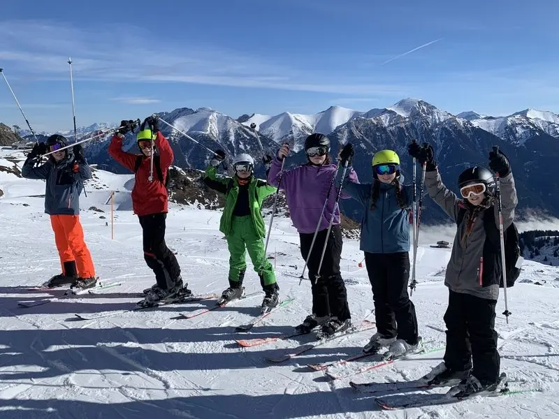 Sechs Jugendliche auf Skiern recken die Stöcke vor verschneiter Bergkulisse in die Höhe