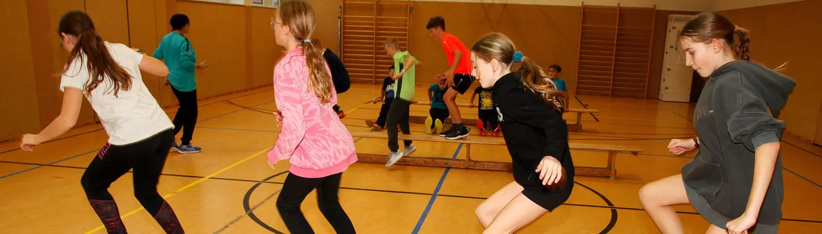 Schülerinnen und Schüler laufen und springen in einer Sporthalle mit aufgestellten Bänken