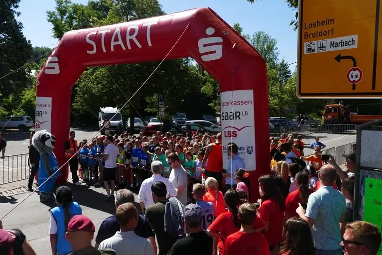 Viele Läuferinnen und Läufer mit Startnummern unter einem aufblasbaren Starttor „Start“, davor Zuschauende