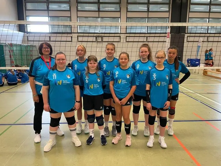 Volleyballmannschaft in türkisfarbenen Trikots mit einer erwachsenen Person vor dem Netz in einer Sporthalle