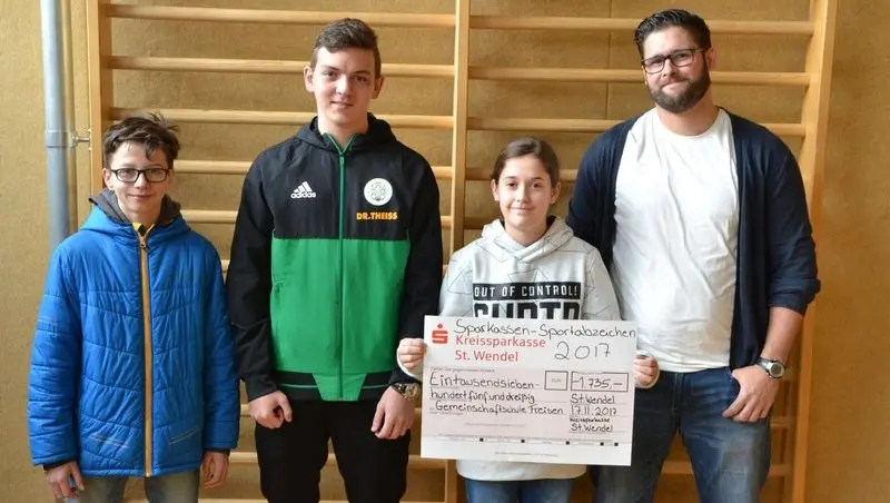 Drei Jugendliche und ein Erwachsener vor einer Sprossenwand mit Spendenscheck der Kreissparkasse über 1735 Euro