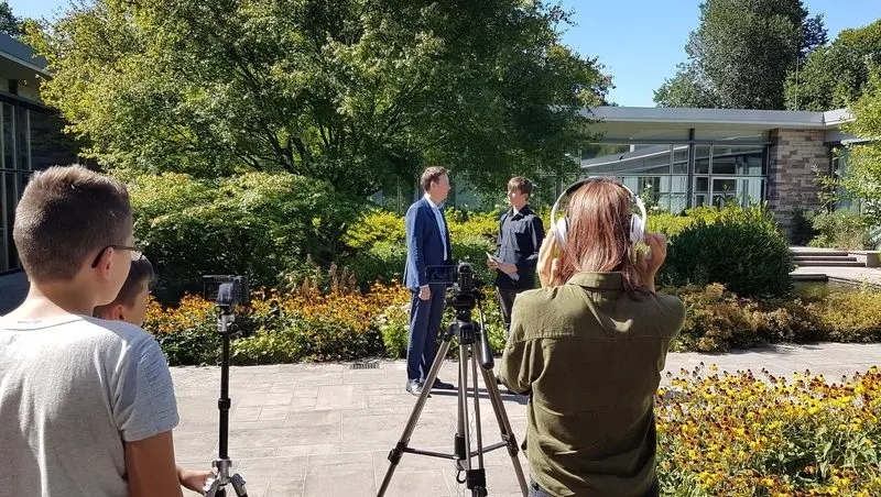 Jugendlicher interviewt einen Mann im Anzug im Garten, im Vordergrund Jugendliche mit Kamera auf Stativ und Kopfhörern