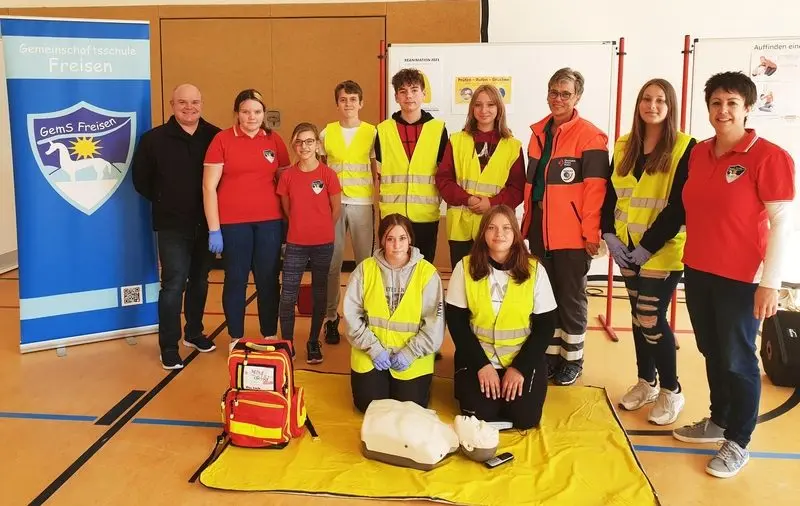 Gruppenbild des Schulsanitätsdienstes in Warnwesten mit Übungspuppe und Notfallrucksack in der Turnhalle