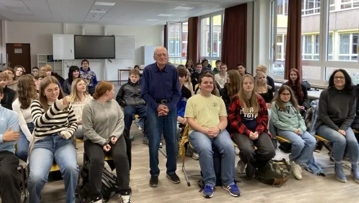 Mann mit weißem Haar steht zwischen sitzenden Schülerinnen und Schülern im Klassenraum