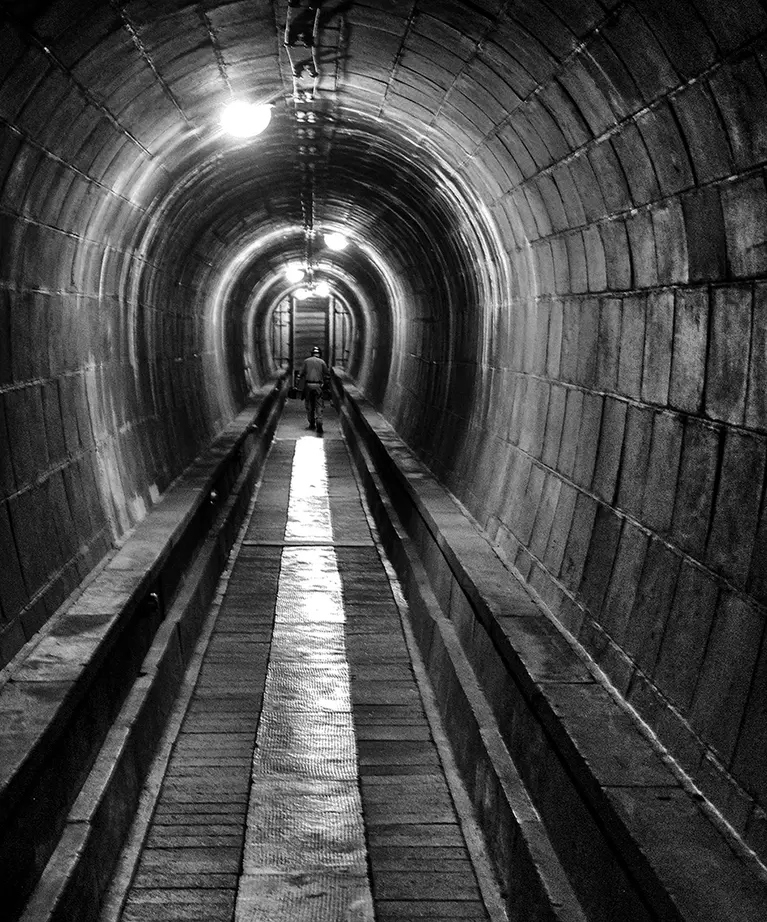 Schwarz-Weiß-Foto: langer gemauerter Tunnel mit Lichtern und einer Person in der Ferne