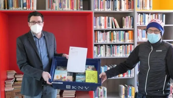 Zwei Männer mit Masken übergeben eine Kiste mit Büchern vor Bücherregalen der Schülerbibliothek