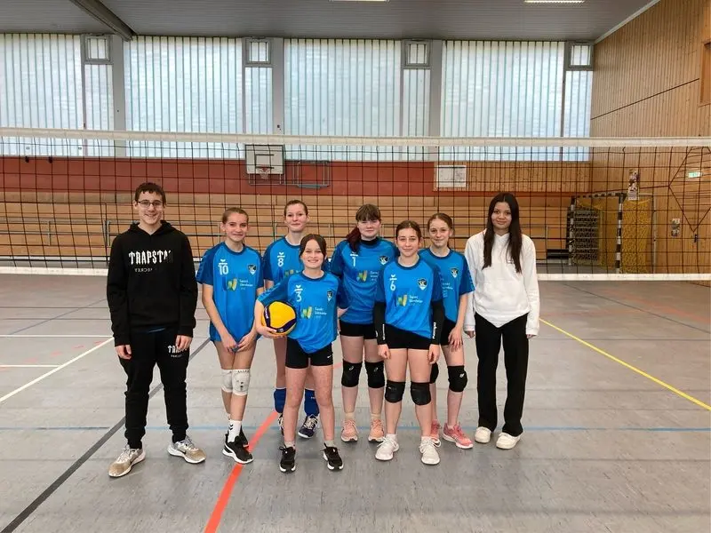 Mädchen-Volleyballmannschaft in blauen Trikots mit Ball in der Sporthalle, daneben zwei Jugendliche