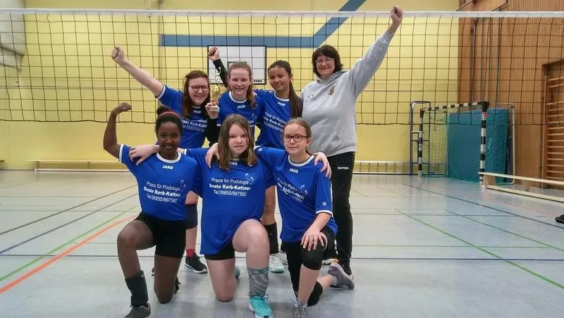 Volleyballerinnen in blauen Trikots jubeln mit Pokal und Trainerin in der Sporthalle