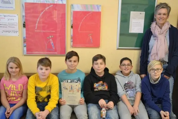 Sechs Kinder sitzen auf einer Bank, eines hält eine Urkunde, dahinter eine Frau