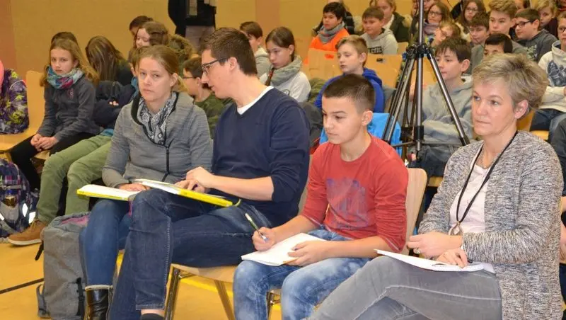 Vier Personen mit Bewertungsbögen in der ersten Reihe, dahinter zuhörende Schülerinnen und Schüler