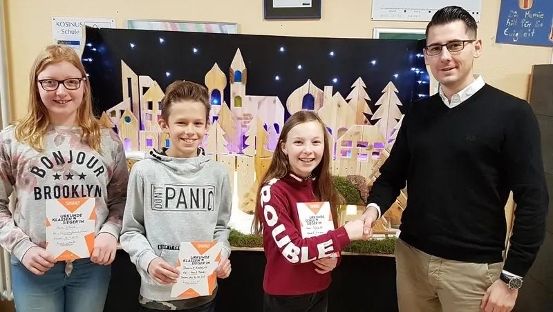 Drei Kinder mit Urkunden vor einer beleuchteten Holzstadt, ein Mann gratuliert der Schulsiegerin per Handschlag