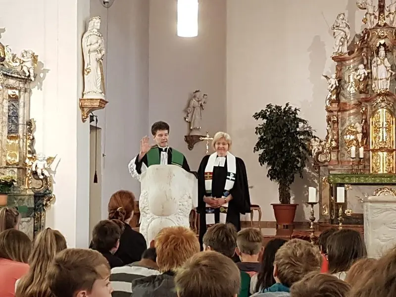 Geistlicher und Pfarrerin im Talar am Altar einer Kirche, er hebt segnend die Hand, davor Schülerinnen und Schüler