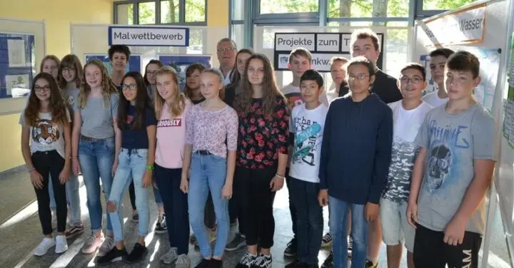 Schülergruppe mit Erwachsenen in einer Ausstellung vor Tafeln, eine mit der Aufschrift „Malwettbewerb“