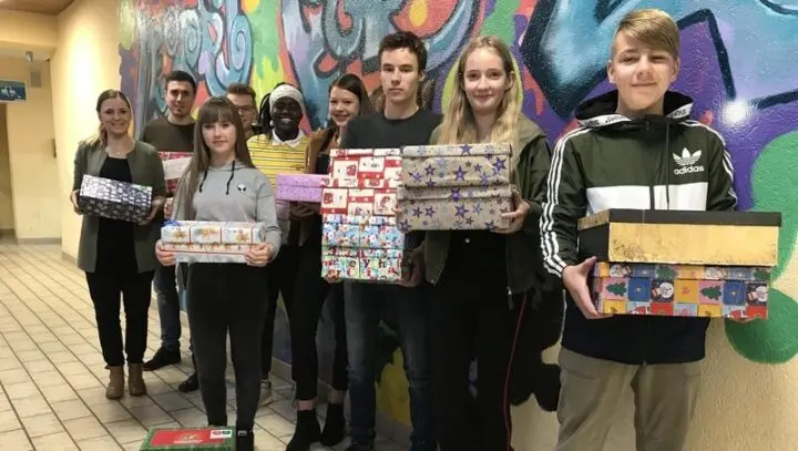 Jugendliche und Erwachsene halten bunt verpackte Schuhkartons vor einer Graffitiwand im Schulflur