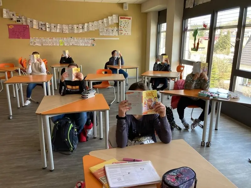 Schülerinnen und Schüler sitzen an Tischen und halten das Buch „Biber undercover“ vor ihr Gesicht