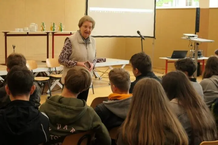 Eine Frau spricht im Saal zu sitzenden Schülerinnen und Schülern, dahinter eine Leinwand