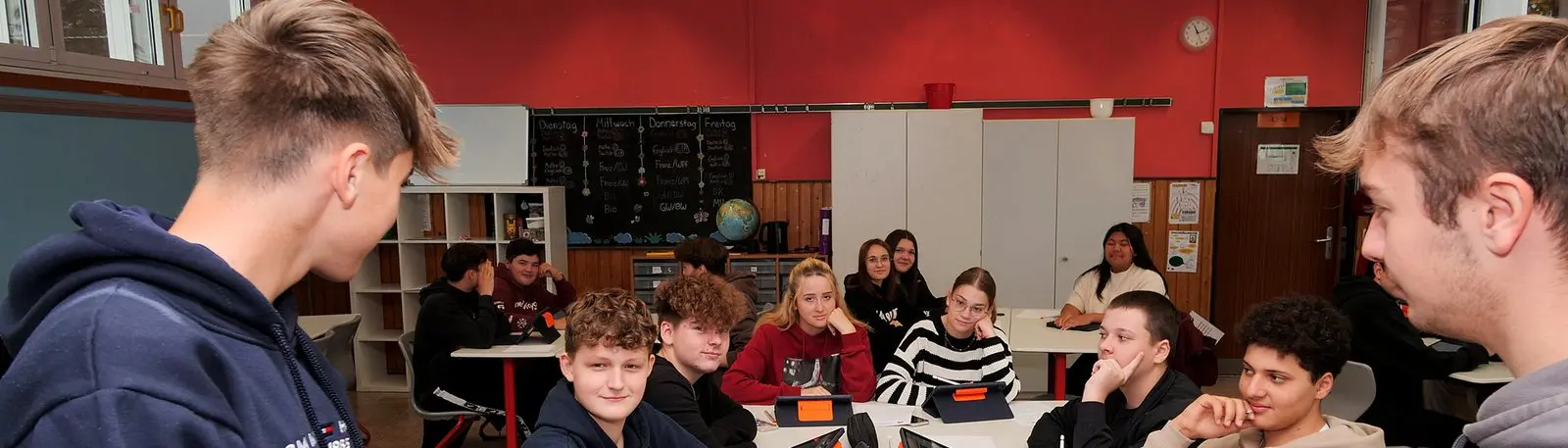 Zwei Jugendliche stehen vor einer Klasse, die an Gruppentischen mit Tablets sitzt