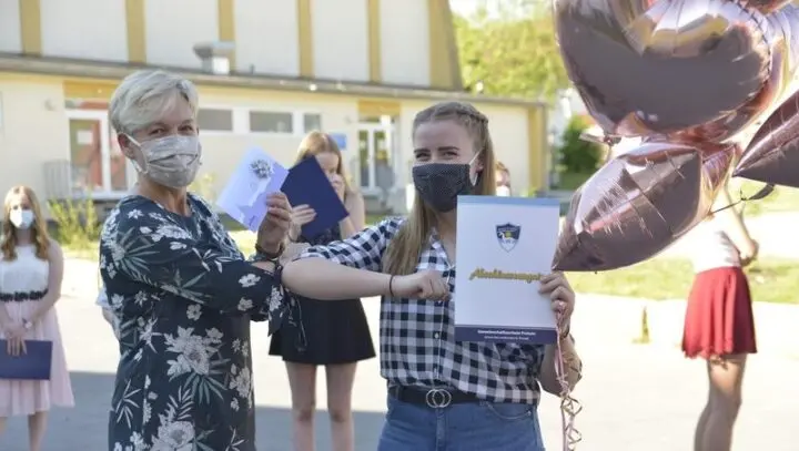 Lehrerin mit Maske übergibt einer Schülerin das Abschlusszeugnis auf dem Schulhof, daneben Luftballons