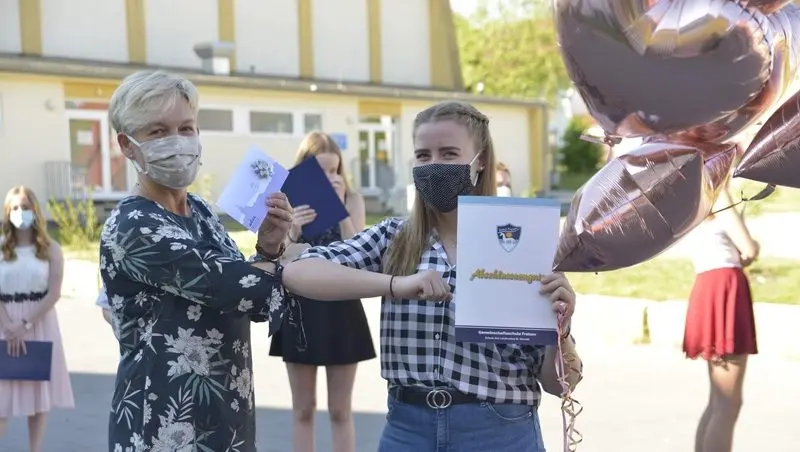 Lehrerin mit Maske übergibt einer Schülerin das Abschlusszeugnis auf dem Schulhof, daneben Luftballons