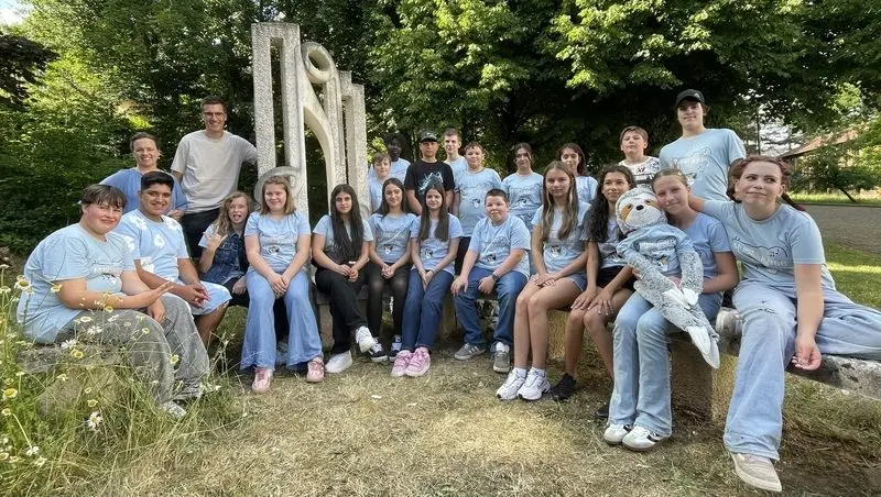 Gruppe von Schülerinnen und Schülern in hellblauen T-Shirts vor einer Steinskulptur, eine hält ein Plüschfaultier