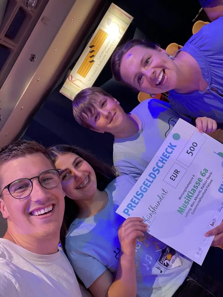 Selfie von zwei Erwachsenen und zwei Kindern der MusiKlasse mit einem Preisgeldscheck über 500 Euro