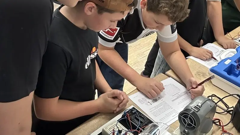 Schüler beugen sich über ein Arbeitsblatt und einen Versuchsaufbau mit Messgerät und Ventilator